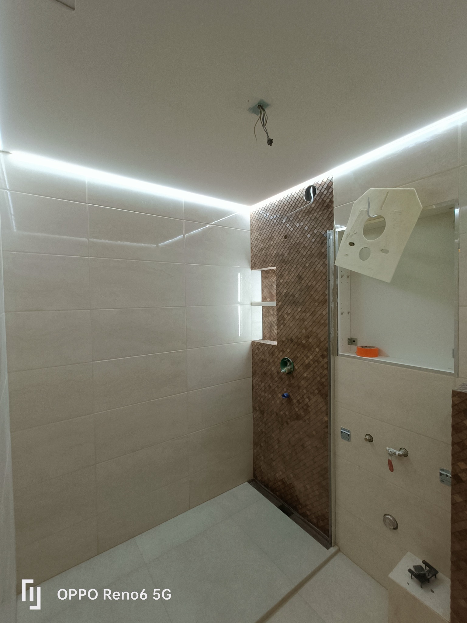 Niedokończona łazienka z beżowymi płytkami ściennymi, dekoracyjną ścianą prysznicową z mozaiką w odcieniach brązu i podświetleniem LED podsufitowym, widoczne niedokończone podłączenia elektryczne...