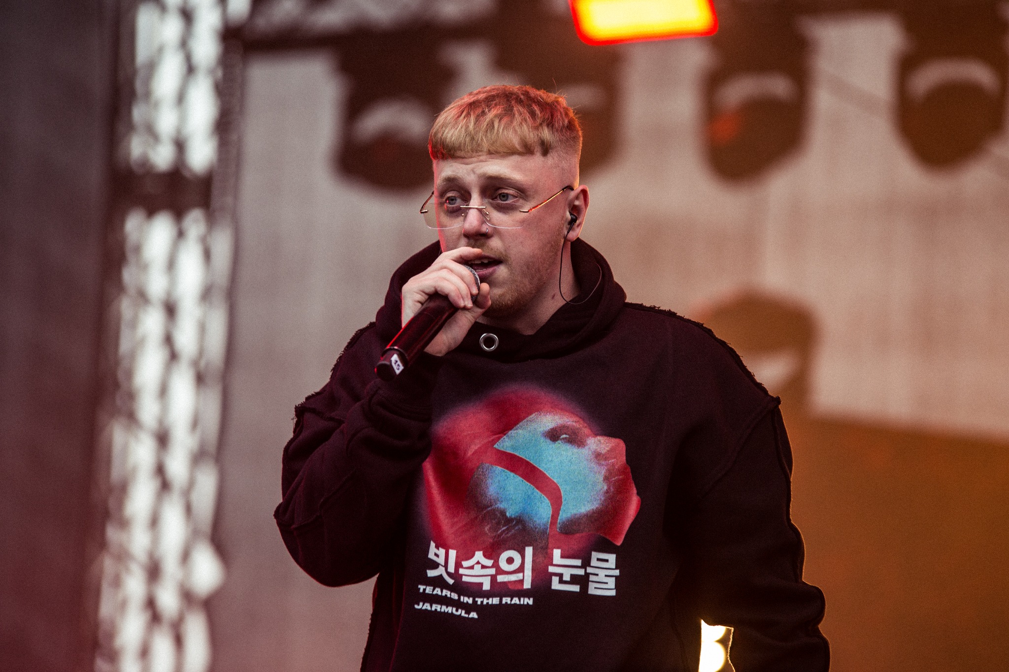 Młody mężczyzna w okularach i bluzie z kapturem, śpiewa do mikrofonu na scenie oświetlonej reflektorami podczas festiwalu muzycznego.