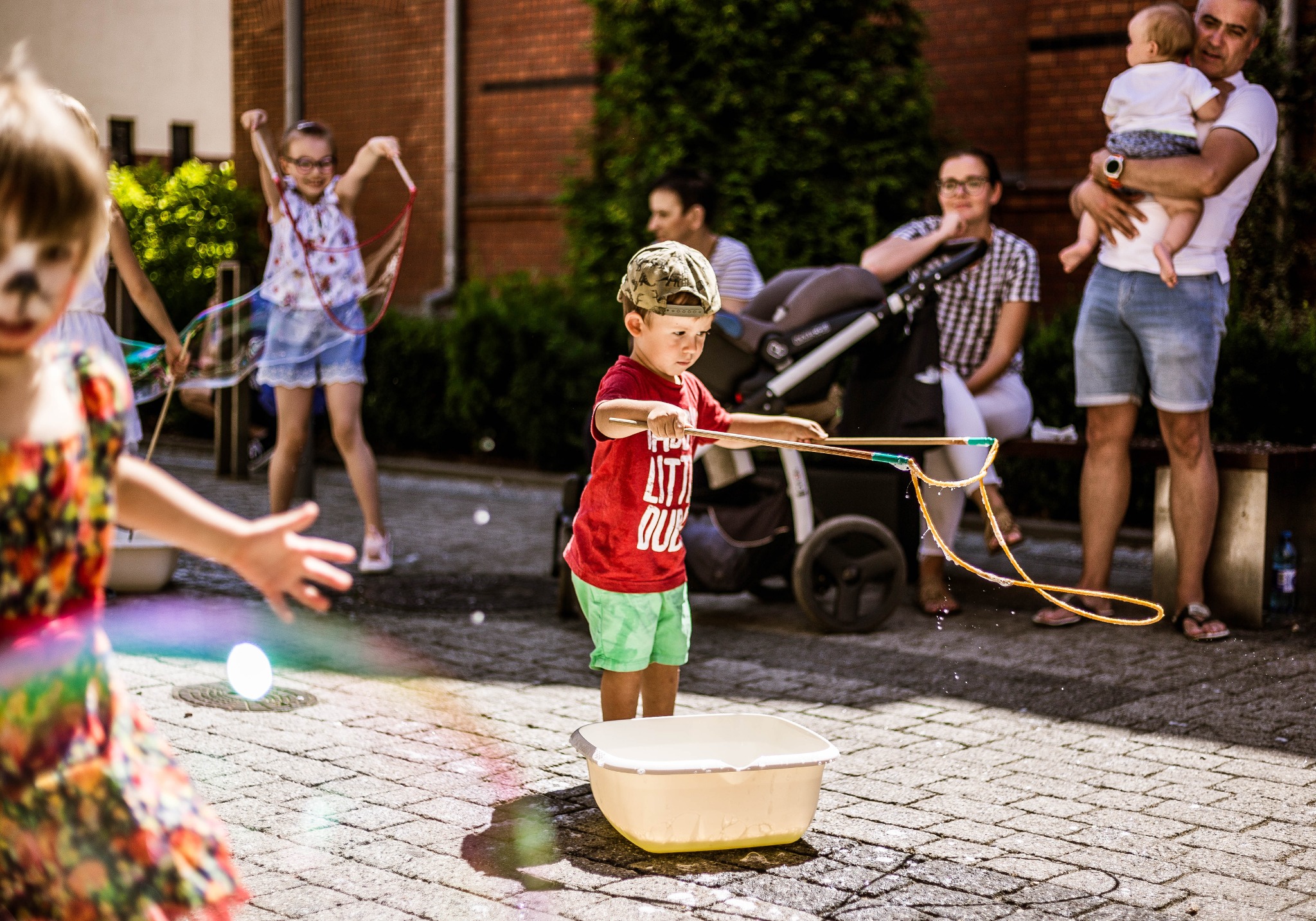 Dzieci puszczają bańki mydlane podczas eventu plenerowego w galerii handlowej, na tle widać spacerujących ludzi i budynek z czerwonej cegły.