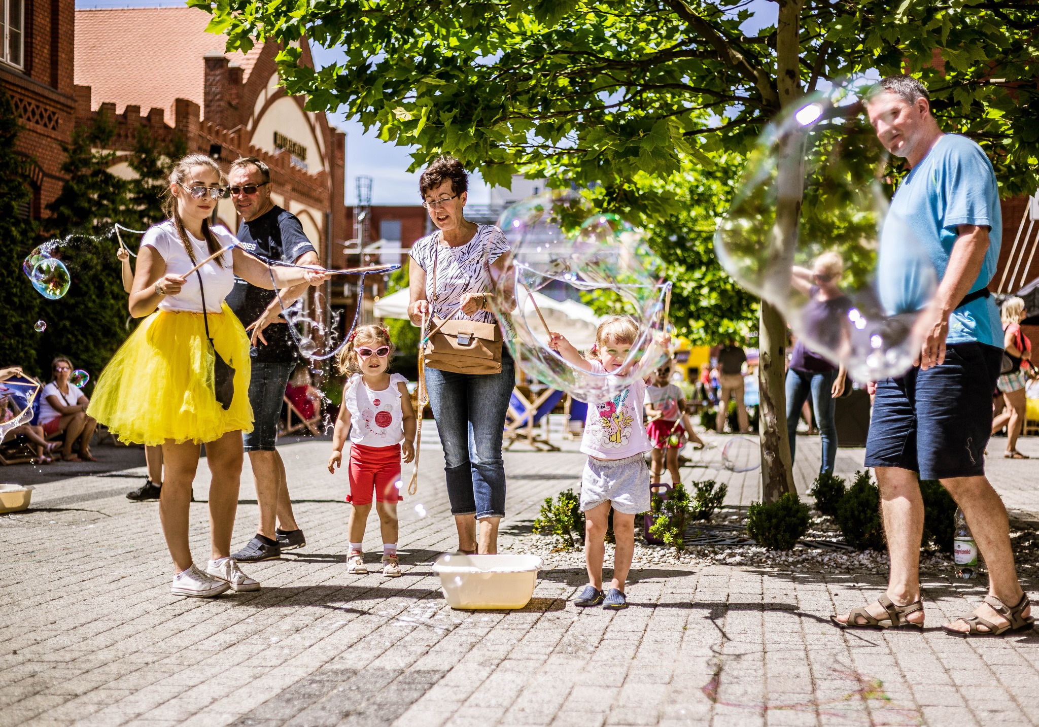 Animatorzy i dzieci puszczają gigantyczne bańki mydlane na zewnątrz, w tle budynki z czerwonej cegły i drzewa.