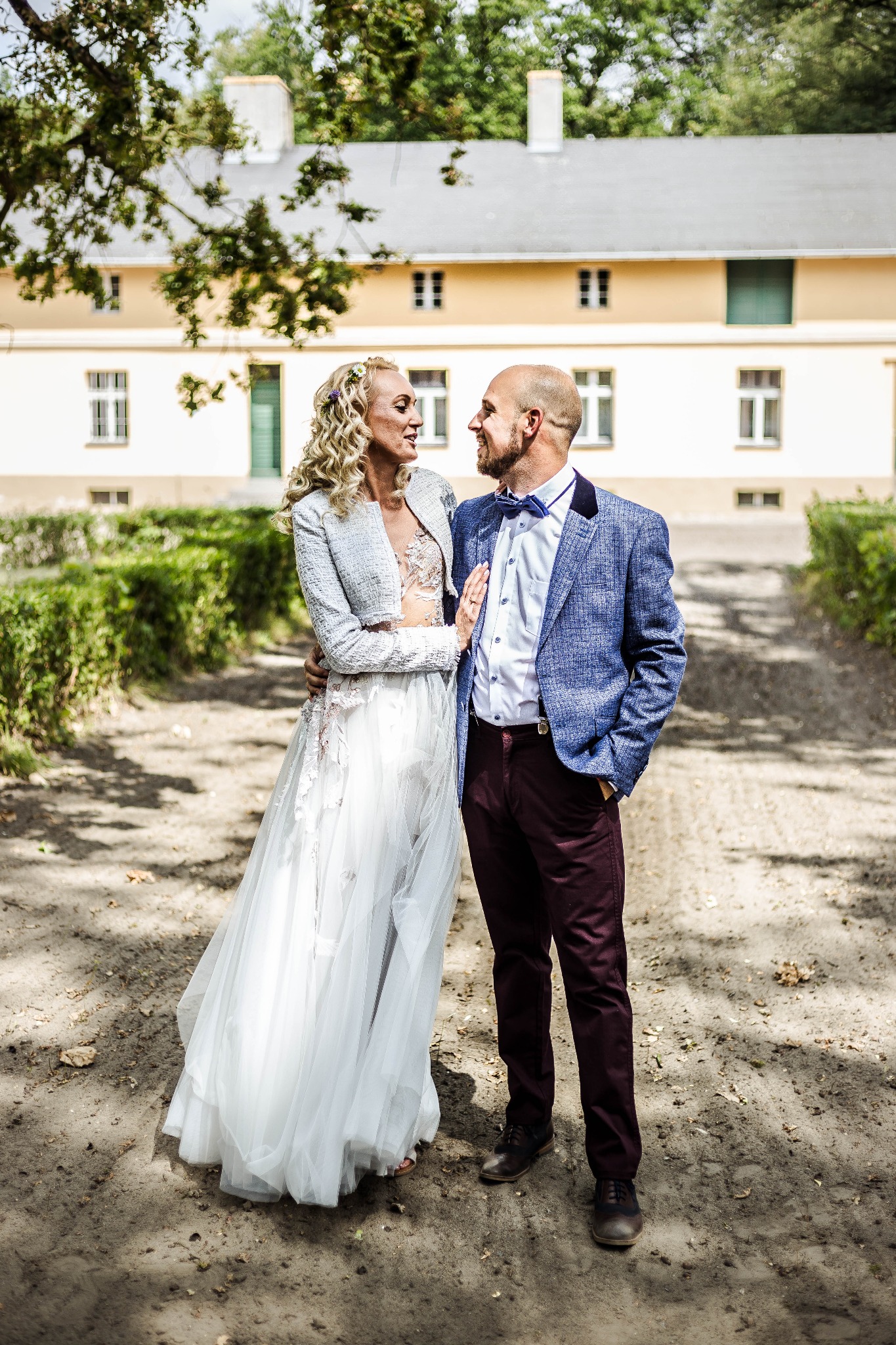 Para młoda w plenerze: Panna młoda w zwiewnej sukni i tweedowej marynarce, pan młody w niebieskiej marynarce w kratę i bordowych spodniach, stoją na tle klasycystycznego budynku z zielonymi...