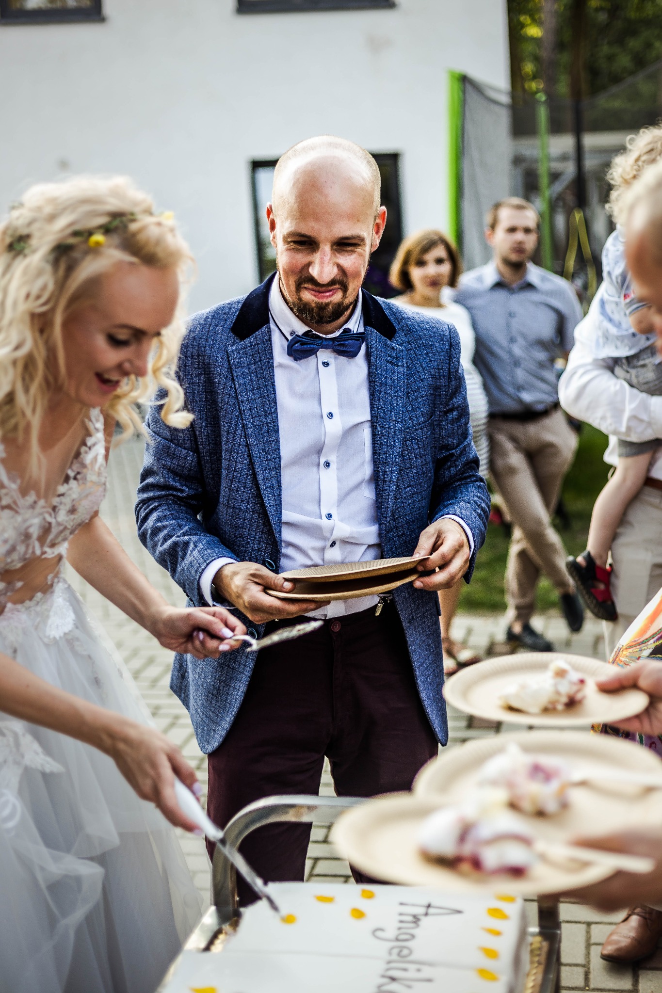 Para młoda kroi tort weselny na zewnątrz; panna młoda w koronkowej sukni, pan młody w niebieskiej marynarce i muszce, goście w tle.