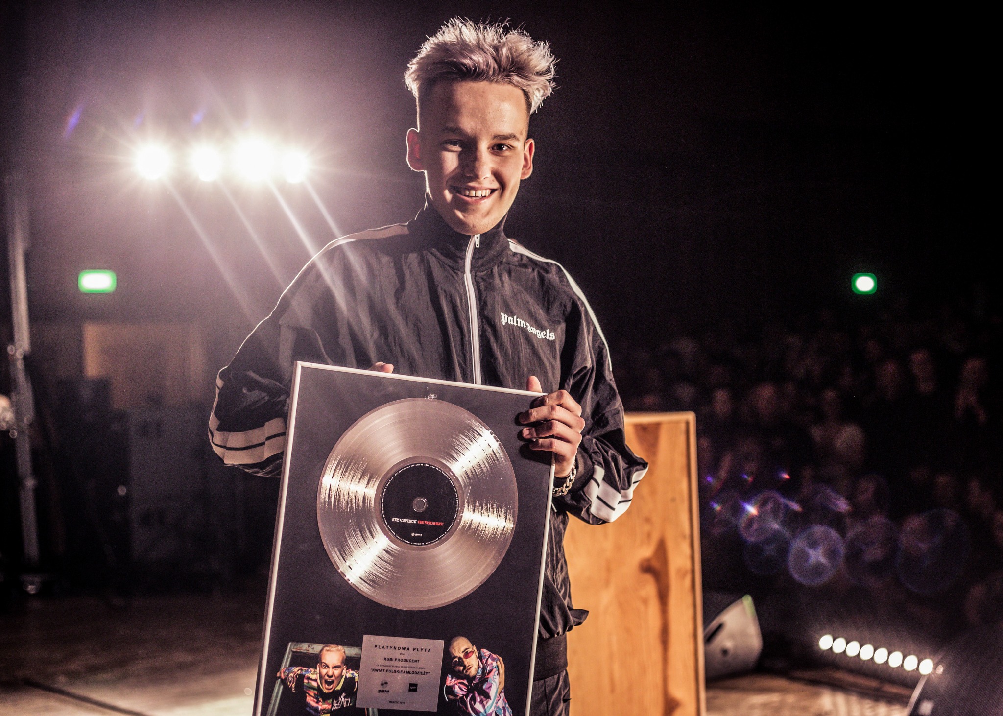 Młody mężczyzna z platynową płytą na tle publiczności i świateł scenicznych, uśmiechnięty, ubrany w czarną kurtkę sportową.