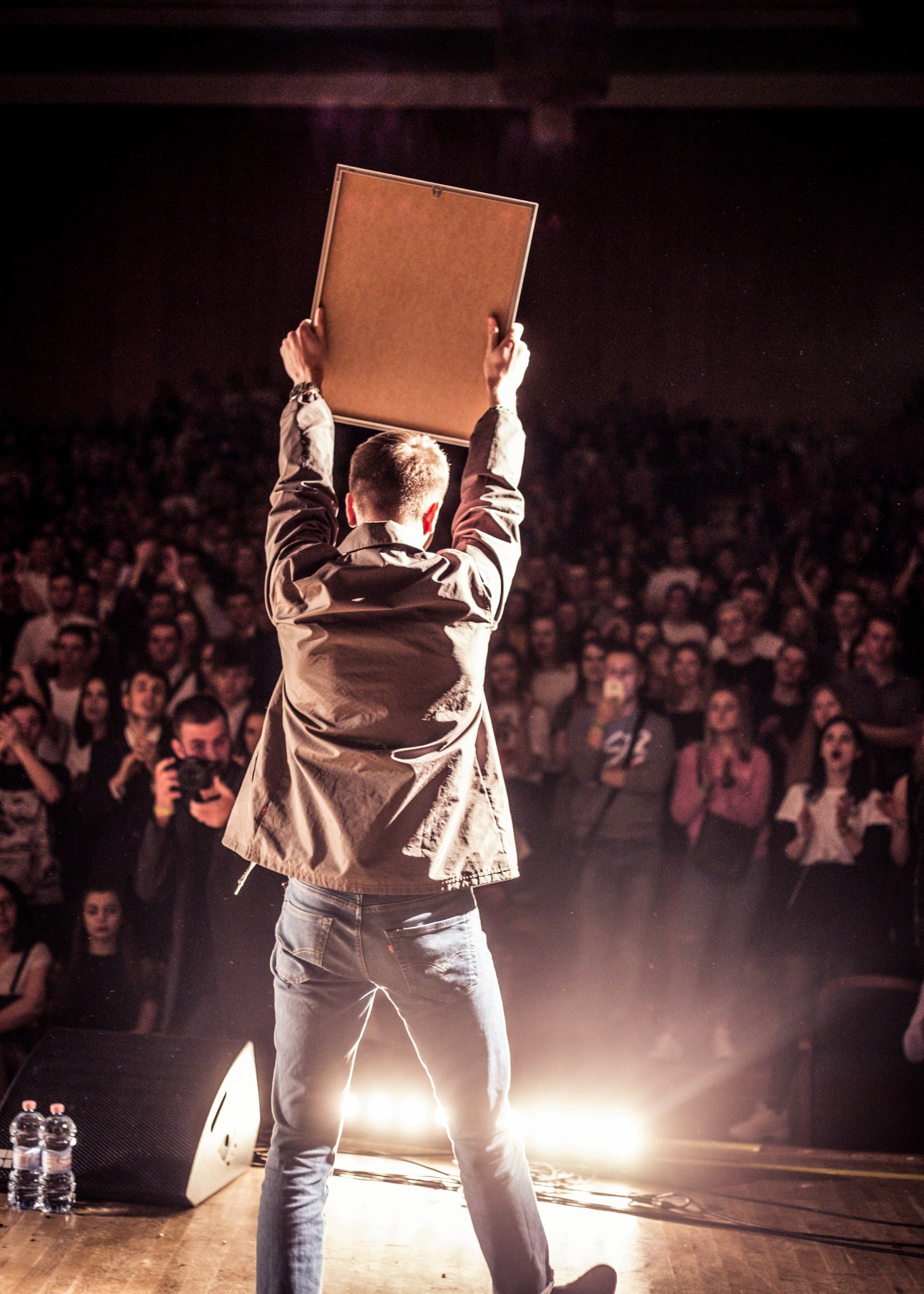 Mężczyzna na scenie, tyłem do widza, trzymający w górze oprawiony obraz na tle rozentuzjazmowanego tłumu podczas koncertu.
