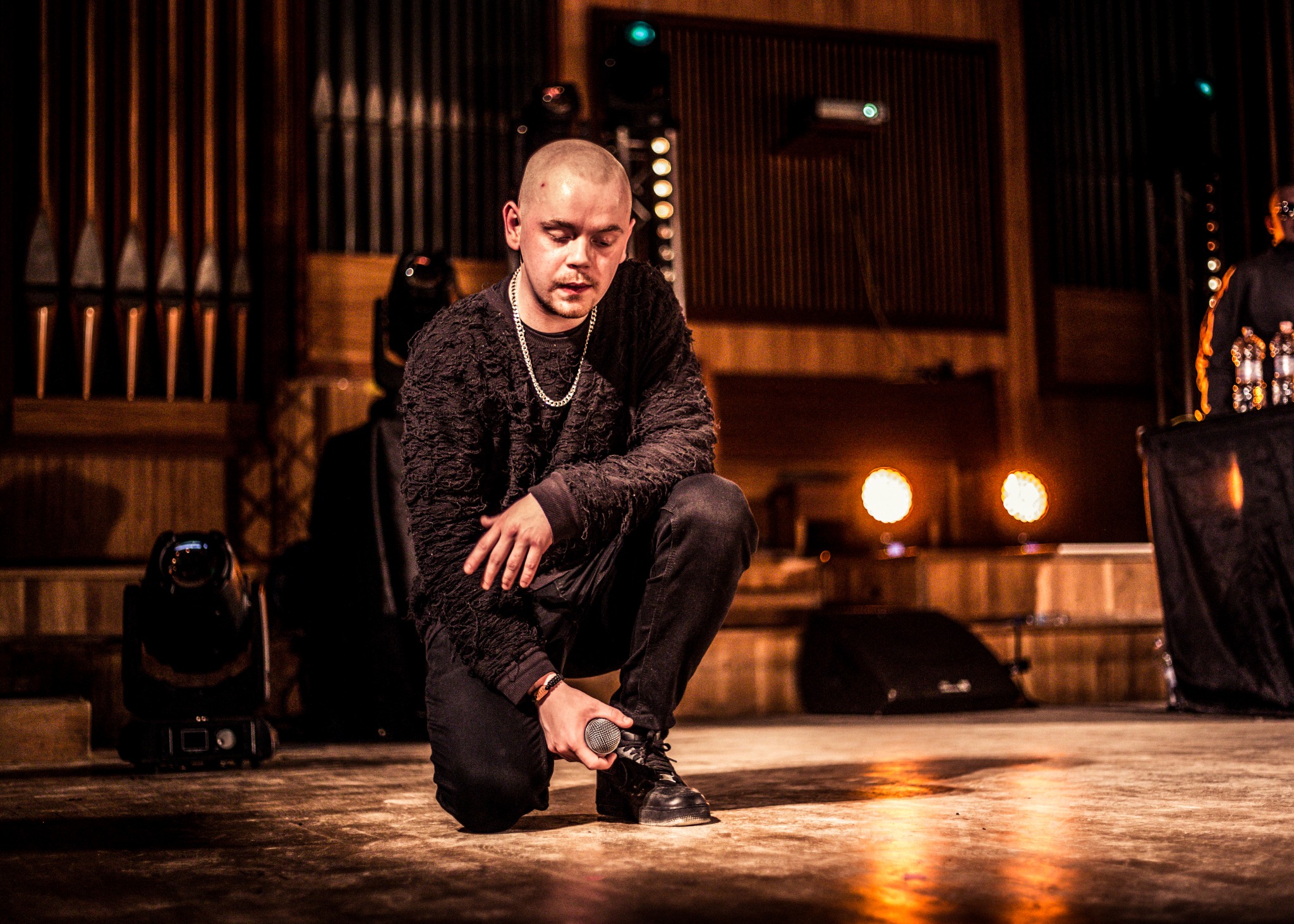 Mężczyzna w czarnym ubraniu klęczy na scenie, trzymając mikrofon w dłoni. W tle oświetlenie sceniczne i elementy dekoracji.