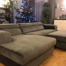 Nowoczesny, szary narożnik z pufą na drewnianej podłodze, w tle choinka z ozdobami i regał z książkami, oświetlenie górne.