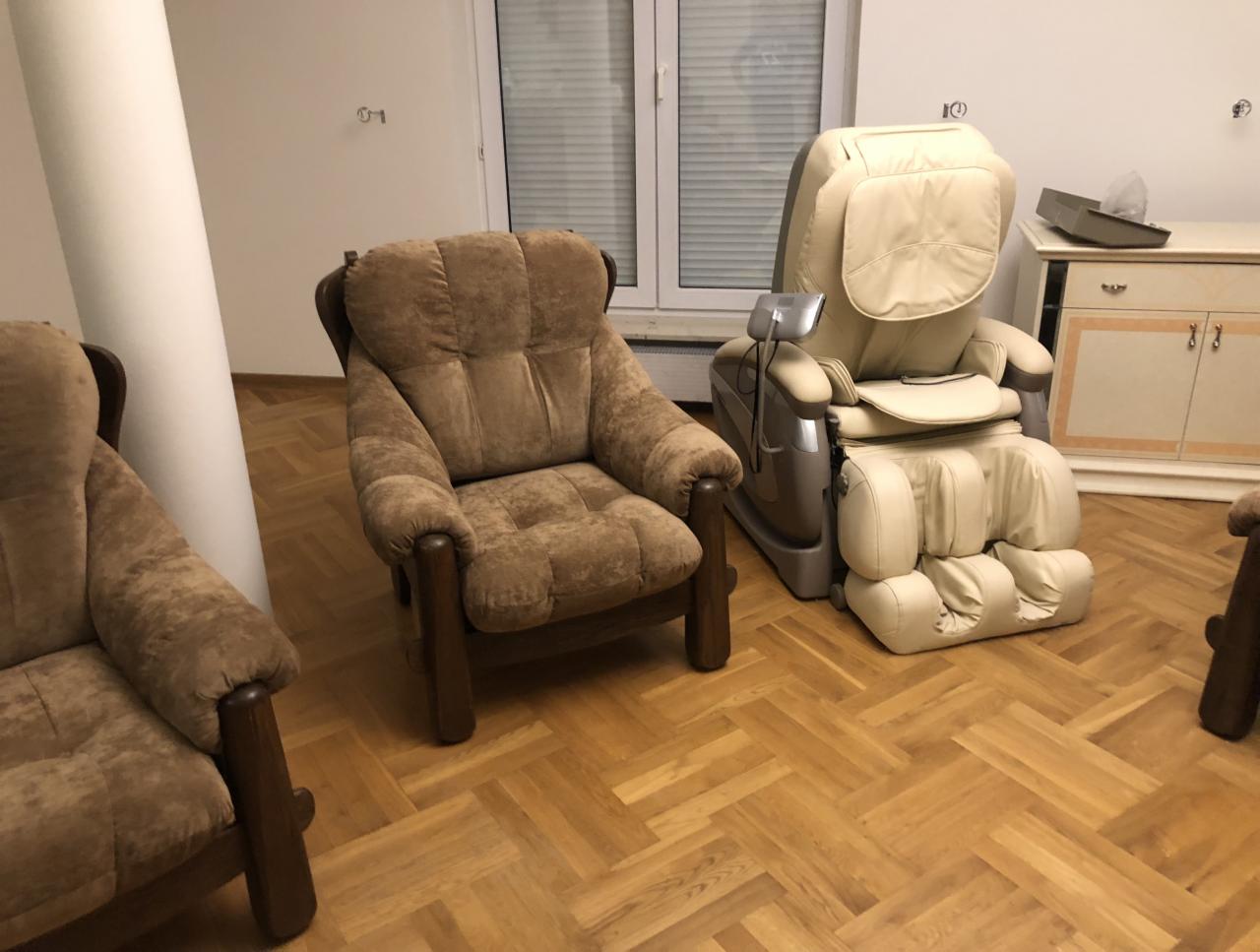 Dwa brązowe, tapicerowane fotele z drewnianymi podłokietnikami oraz beżowy fotel masujący na parkiecie w jasnym pokoju.