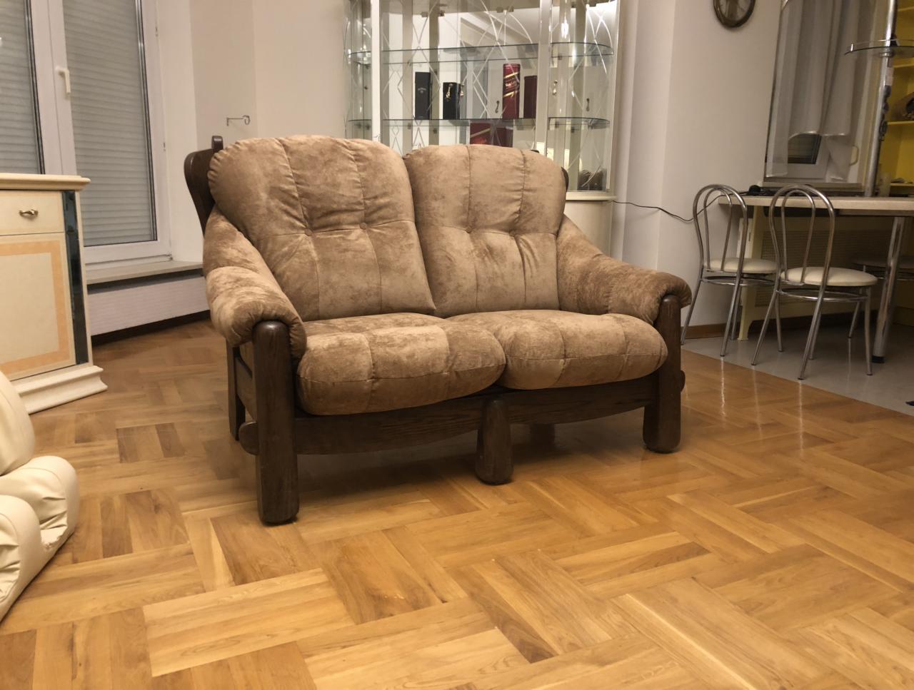 Dwuosobowa kanapa z brązową tapicerką i ciemnobrązowymi drewnianymi nóżkami na parkiecie w jodełkę. W tle widoczny przeszklony regał i zestaw stół z krzesłami.