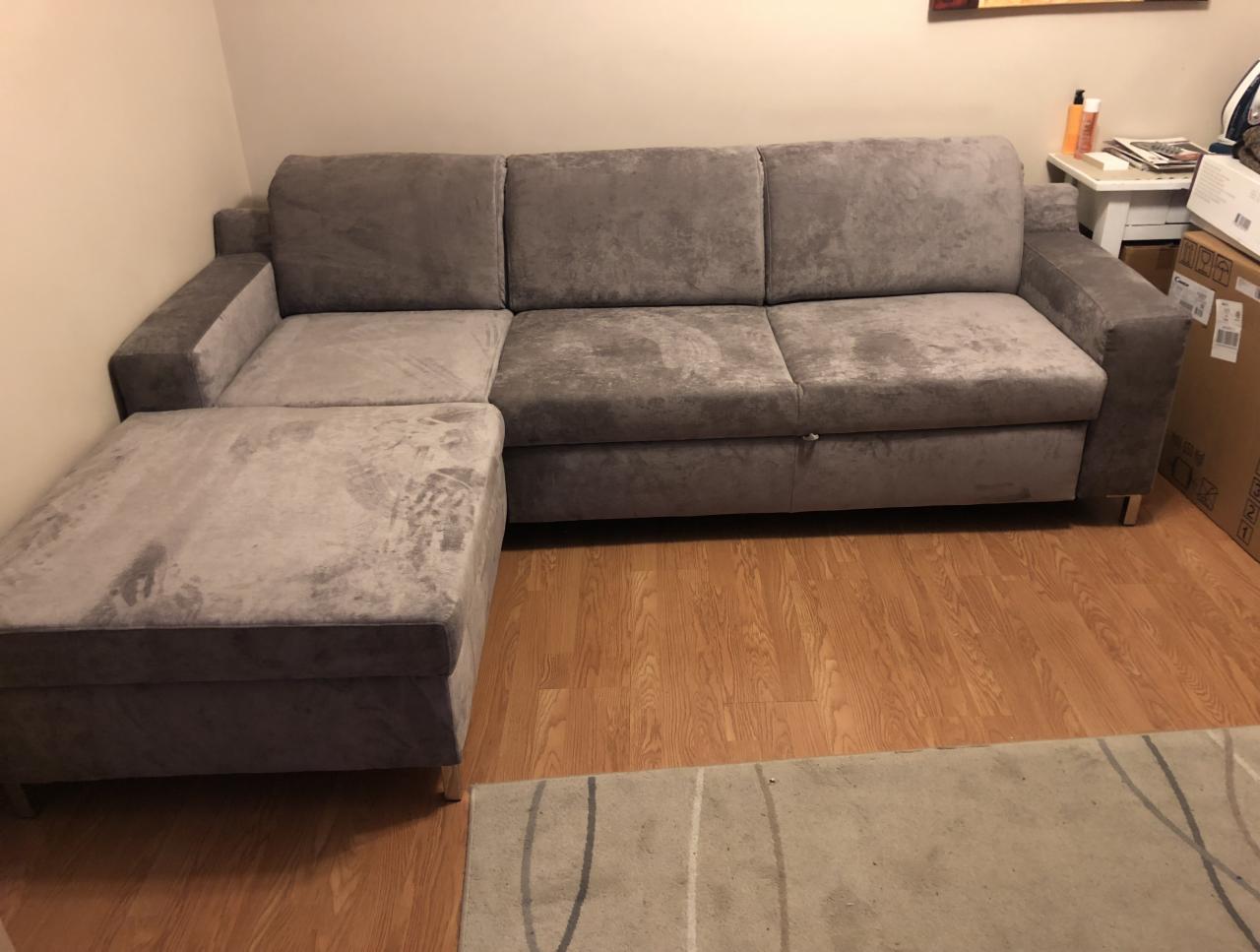 Szary narożnik z funkcją spania i pufą w pokoju z panelami podłogowymi, widoczne ślady użytkowania na tapicerce.