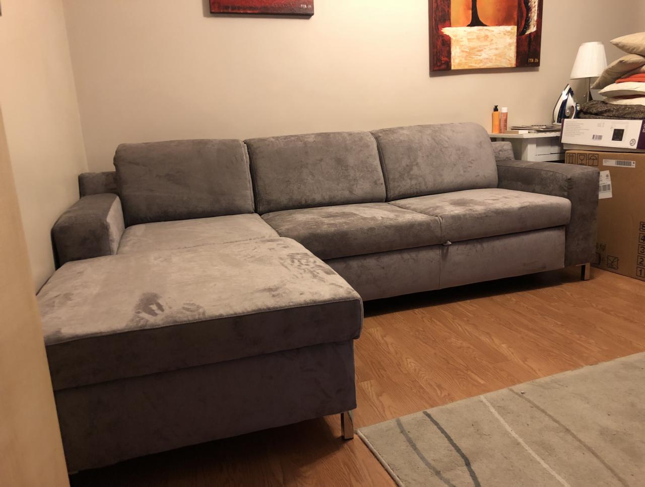 Szara, narożna sofa z podnóżkiem na metalowych nóżkach, widoczna na tle jasnej ściany i drewnianej podłogi, z obrazem na ścianie w tle.