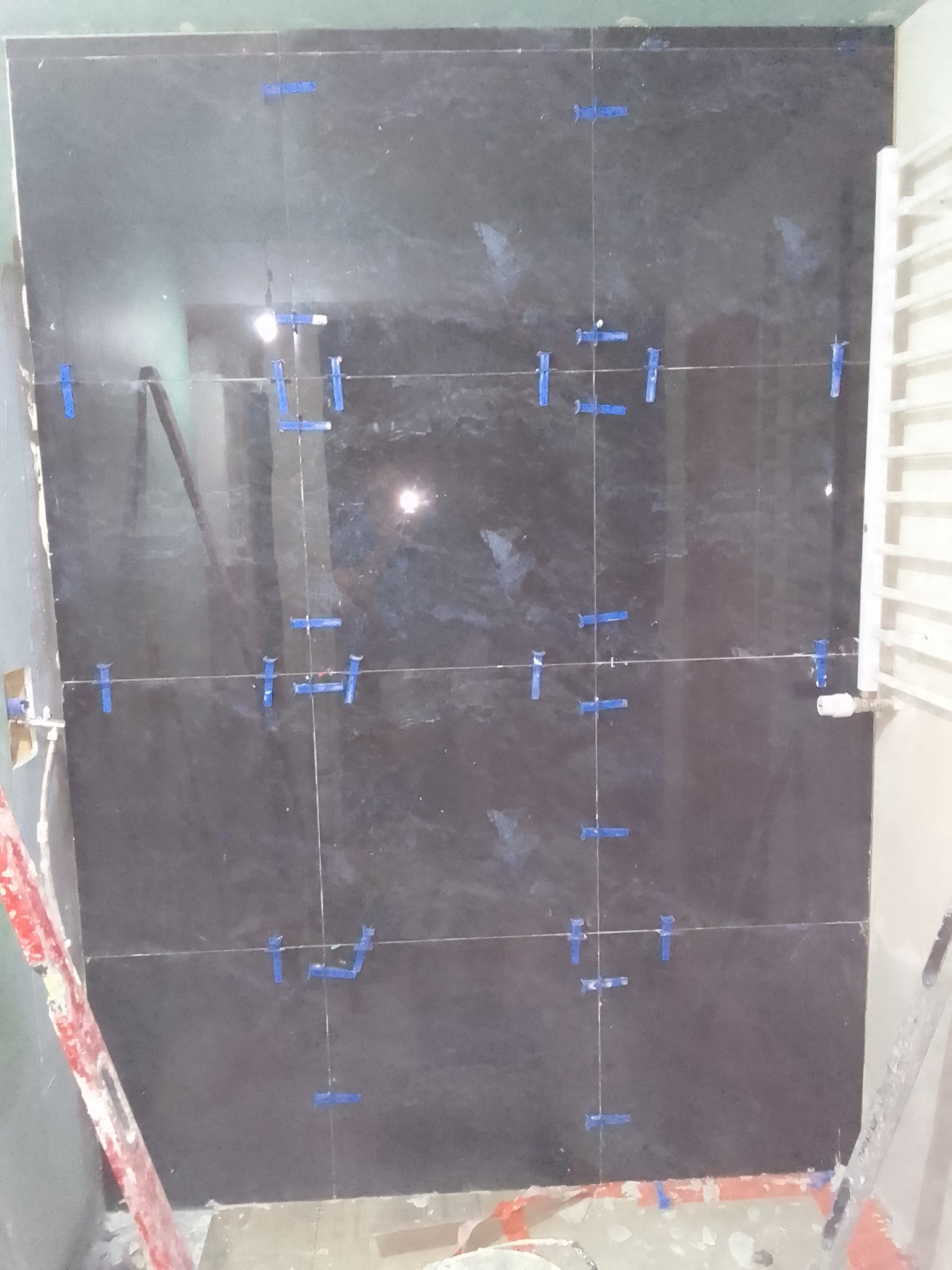 Ciemne płytki na ścianie z niebieskimi krzyżykami dystansowymi, widoczny grzejnik drabinkowy i fragment drabiny.