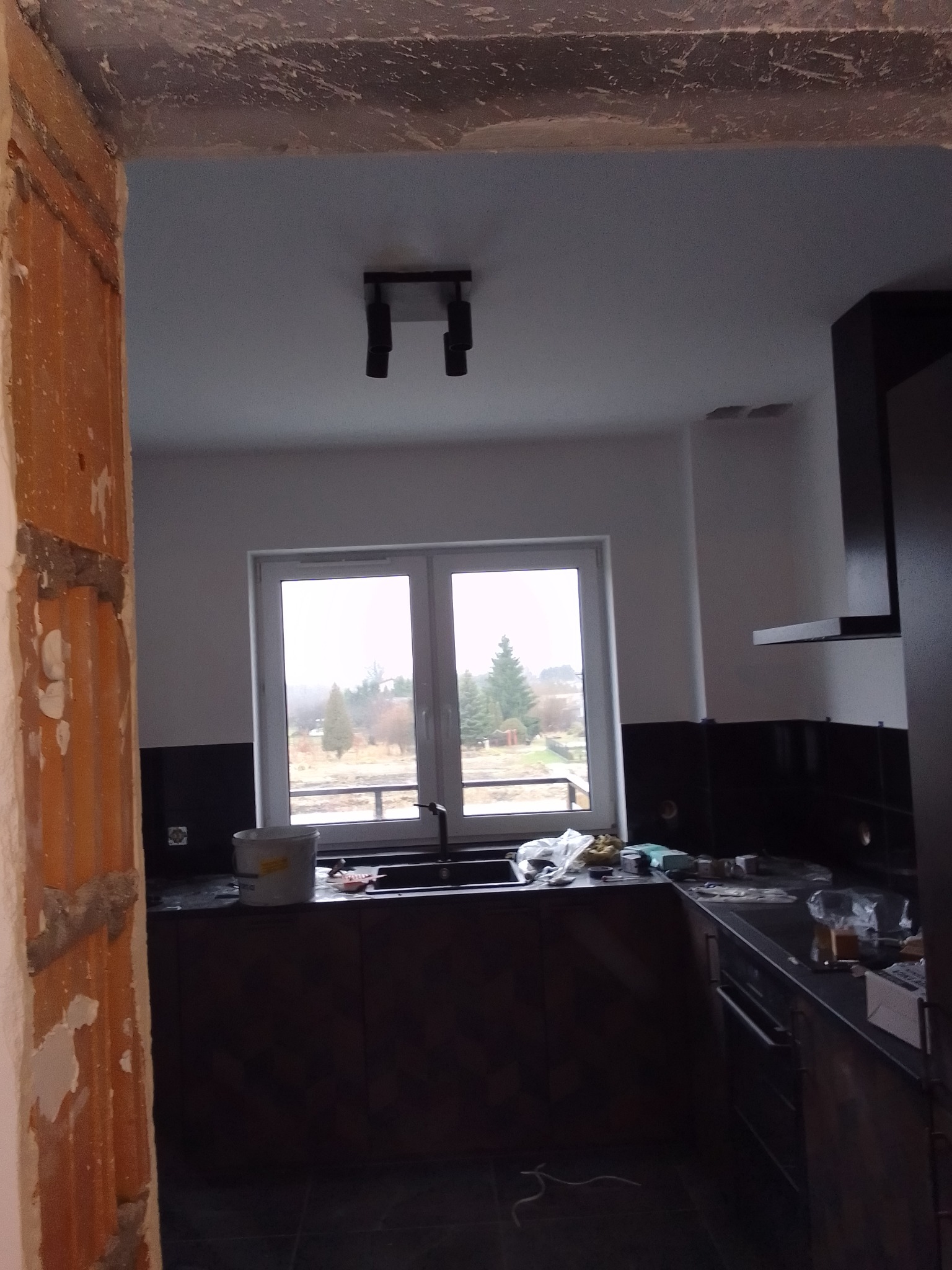 Wnętrze kuchni w trakcie prac wykończeniowych; widoczna surowa ściana z cegły, okno z widokiem na zewnątrz, zabudowa kuchenna z ciemnymi frontami oraz białe ściany z czarnymi płytkami między szafkami.
