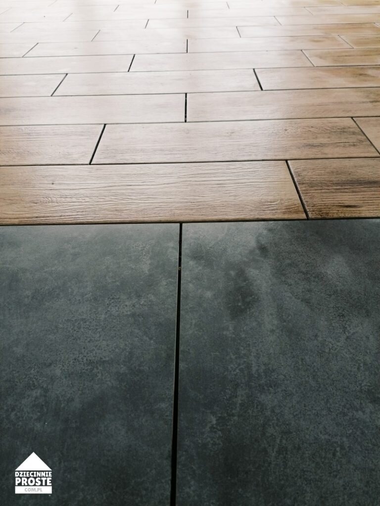Połączenie płytek podłogowych: imitacja drewna ułożona w cegiełkę i ciemnoszara płytka o fakturze betonu, widoczny fragment logo firmy w lewym dolnym rogu.
