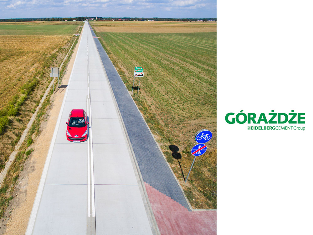 Widok z drona na prostą, betonową drogę przez pola, z czerwonym samochodem osobowym jadącym w kierunku horyzontu. Po prawej stronie drogi, ścieżka rowerowa z kostki brukowej i znaki drogowe.