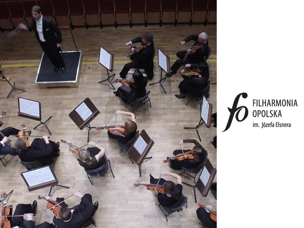 Widok z góry na orkiestrę smyczkową podczas występu; dyrygent stoi na podwyższeniu.