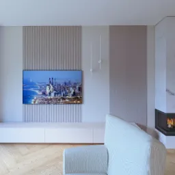 Minimalistyczny salon z kominkiem obudowanym marmurem, telewizorem z widokiem na Dubaj i jasną podłogą z parkietu. Widoczny fragment sofy.