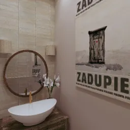 Beżowa łazienka z okrągłym lustrem, umywalką na drewnianej szafce i plakatem „Zadupie”. Na ścianie kafelki imitujące cegłę i dwa kinkiety z abażurami.