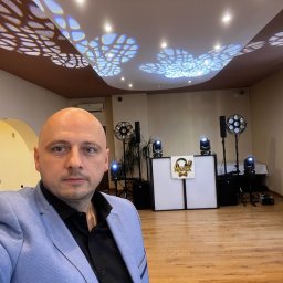 Marian Nowak - DJ w niebieskiej marynarce na tle sprzętu nagłośnieniowego i dekoracji balonowych, oświetlenie z efektem wzorów na suficie.