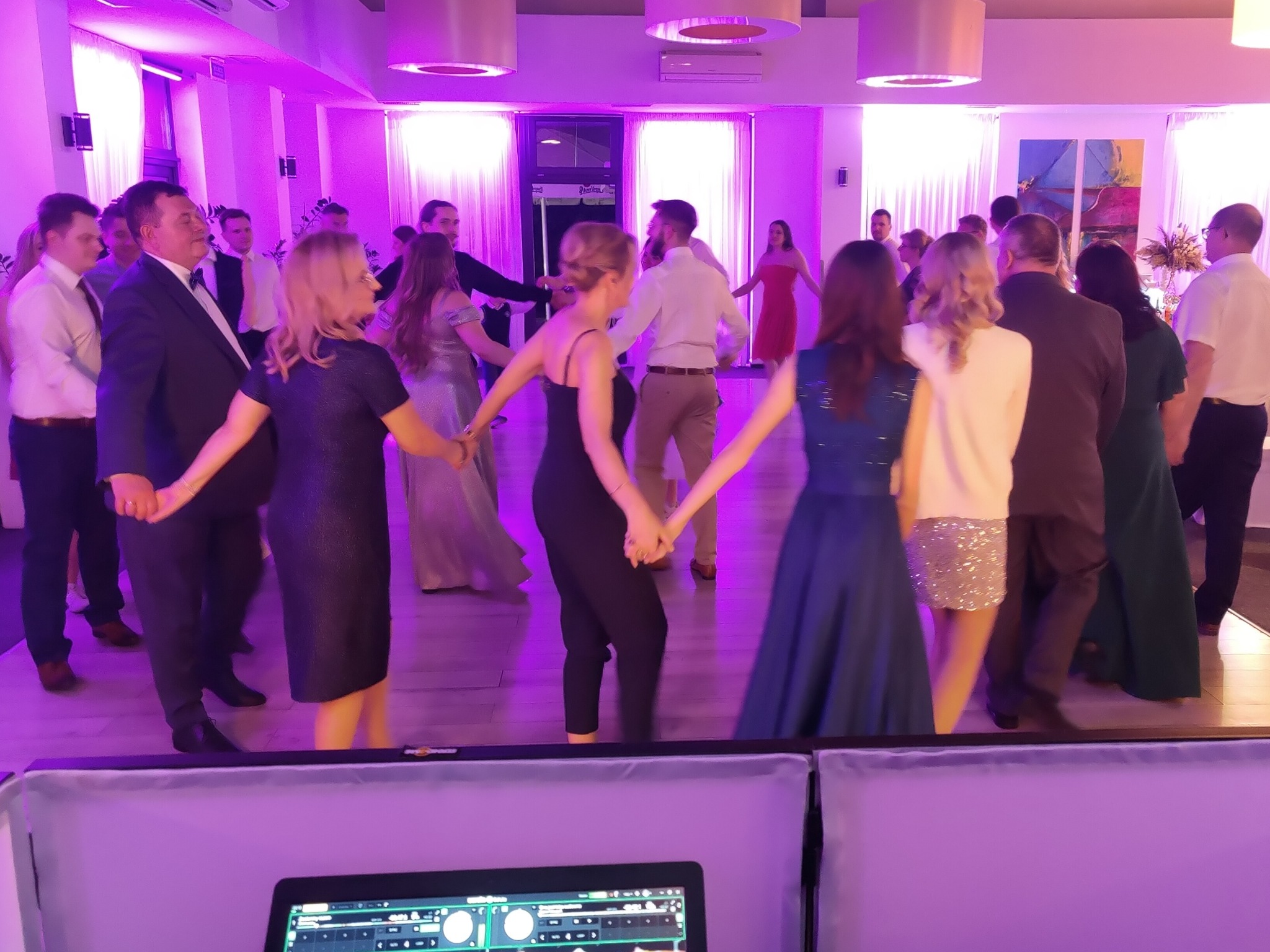 Grupa osób trzymających się za ręce, tańczących w okręgu podczas wydarzenia, oświetlona fioletowym światłem, z widocznym sprzętem DJ-skim na pierwszym planie.