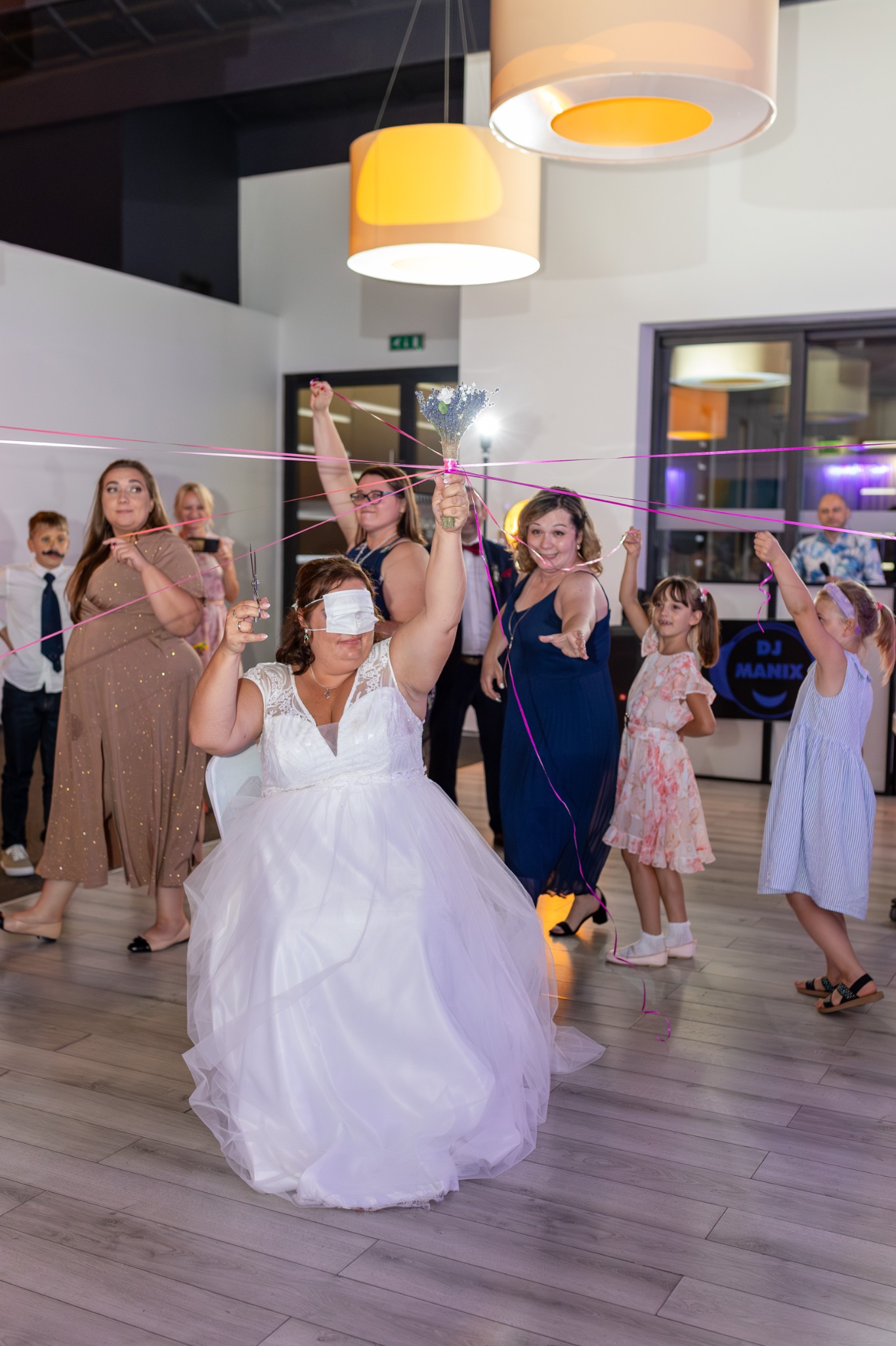 Panna młoda z zawiązanymi oczami, trzymająca nożyczki i bukiet, otoczona wstążkami trzymanymi przez gości podczas tradycyjnej zabawy weselnej.