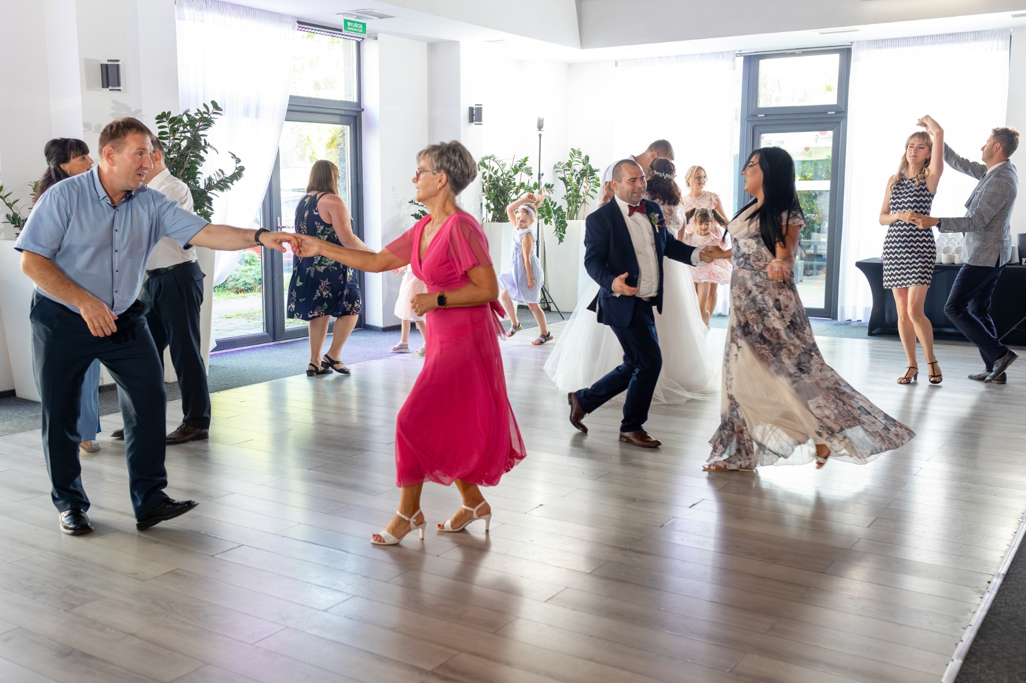 Goście weselni w ruchu podczas tańca, widoczna para młoda w tle, sala z parkietem i roślinami doniczkowymi.