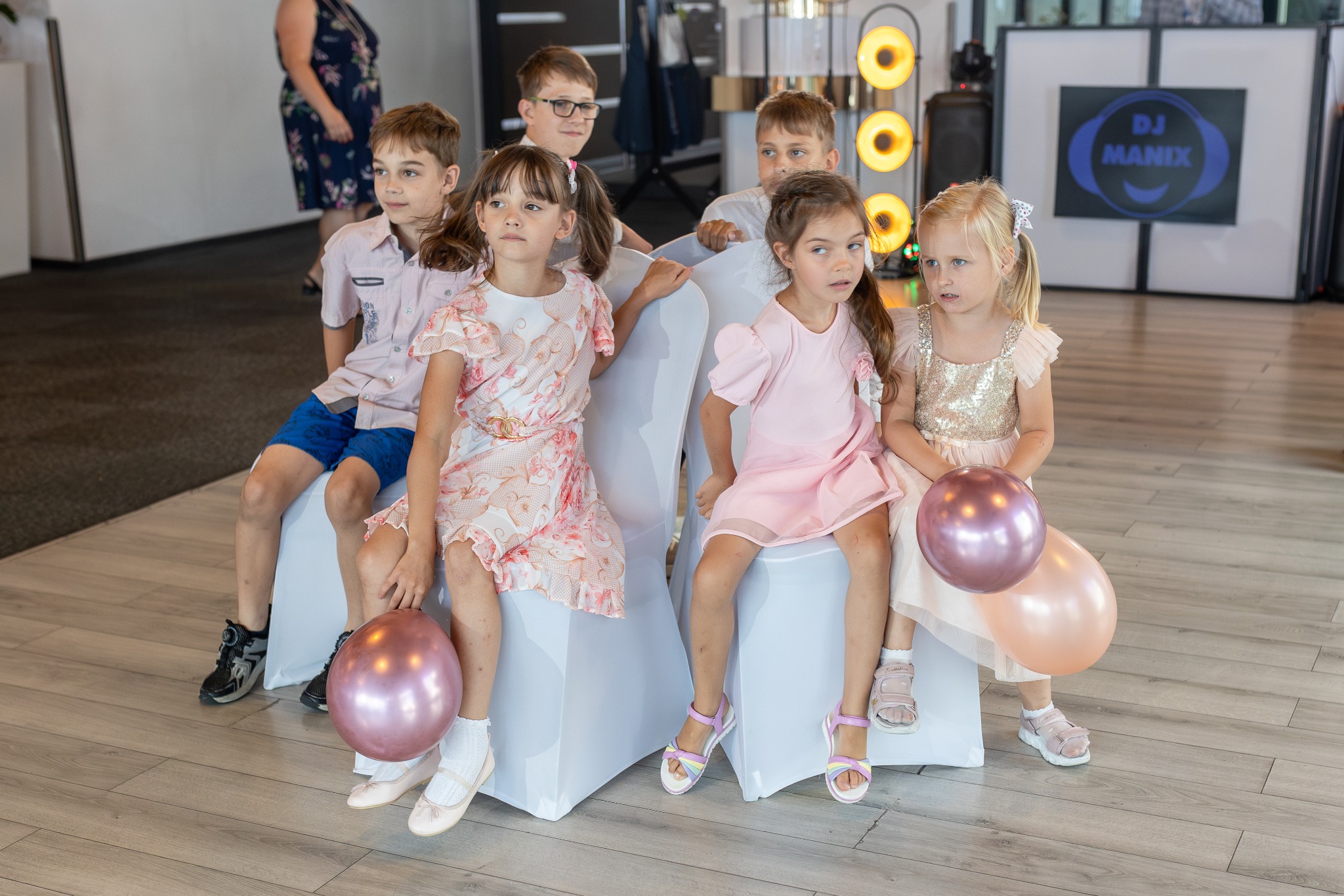 Grupa dzieci w odświętnych strojach siedzi na krzesłach na tle sceny z oświetleniem i logo DJ Manix.