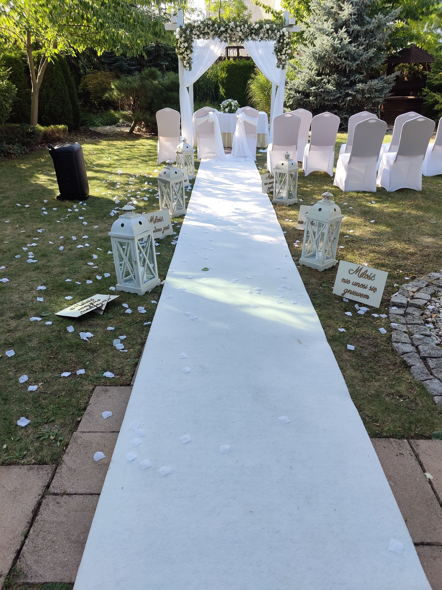 Uroczyste ustawienie plenerowe: biały dywan prowadzi do udekorowanego kwiatami łuku, białe krzesła, lampiony i tabliczki z sentencjami, nagłośnienie po lewej stronie.