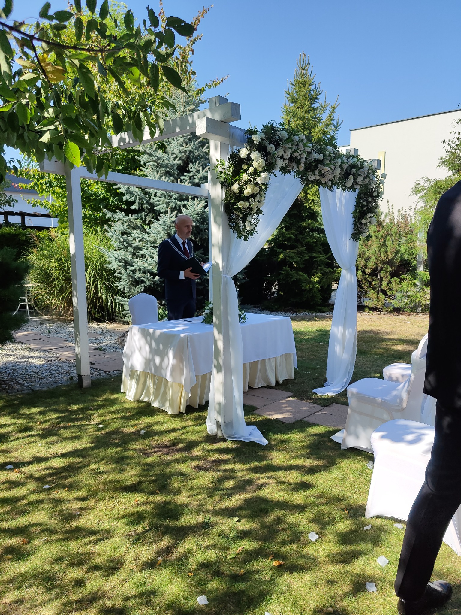 Ceremonia ślubna w plenerze: urzędnik w garniturze odczytuje tekst przy białym stoliku pod ozdobnym baldachimem z kwiatów i białych tkanin, białe krzesła dla gości, trawnik z opadłymi płatkami.