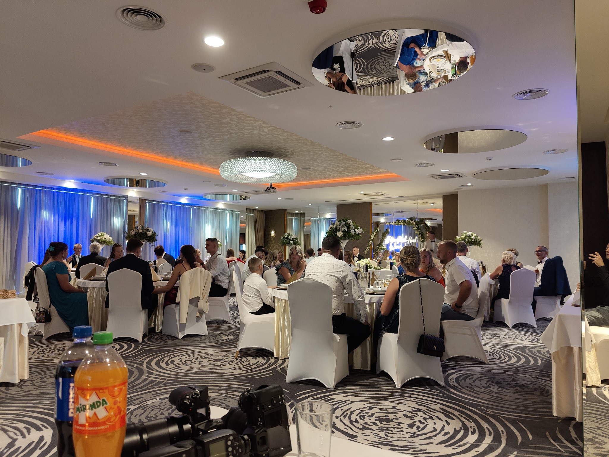 Sala bankietowa w Tarnowskich Górach, przygotowana do przyjęcia weselnego, z okrągłymi stołami nakrytymi białymi obrusami, białymi krzesłami i dekoracjami kwiatowymi, widok zza kamery wideo...