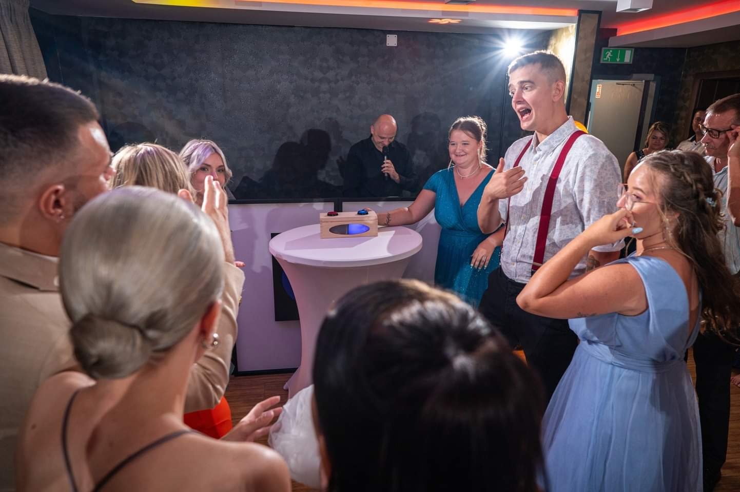 Goście weselni w Tarnowskich Górach obserwują interaktywną zabawę z drewnianym pudełkiem z przyciskami na podświetlonym stoliku, konferansjer z mikrofonem w tle.