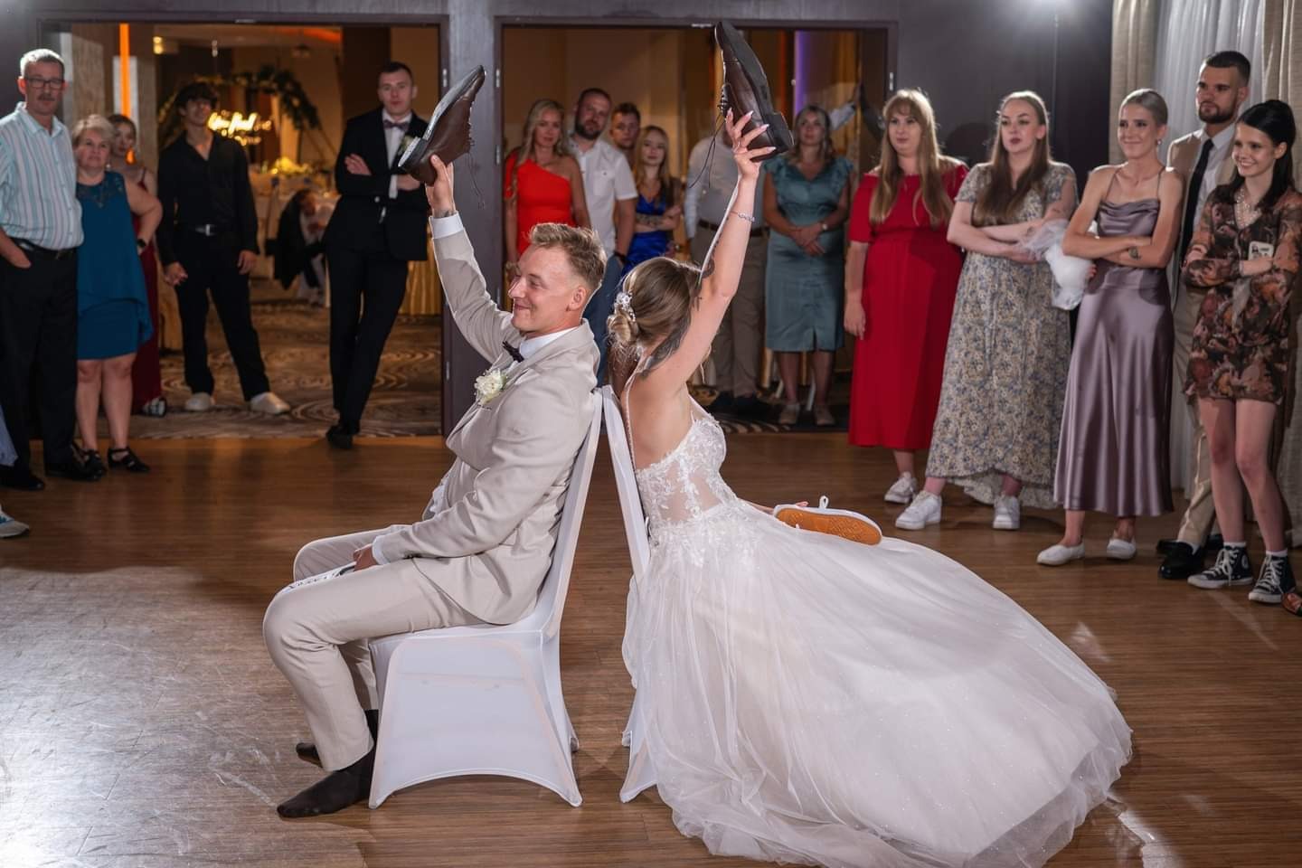 Para młoda siedzi tyłem do siebie na krzesłach, trzymając w górze buty, podczas weselnej zabawy w obecności gości.