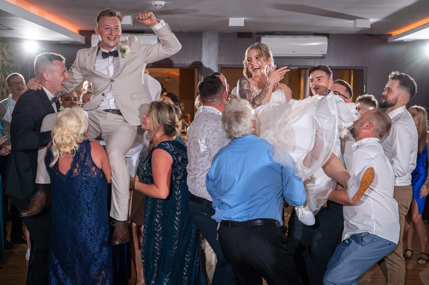 Radosna para młoda niesiona na rękach gości weselnych podczas tradycyjnego oczepin, pan młody w beżowym garniturze z muszką, panna młoda w białej sukni z tatuażami na ramionach, sala weselna...