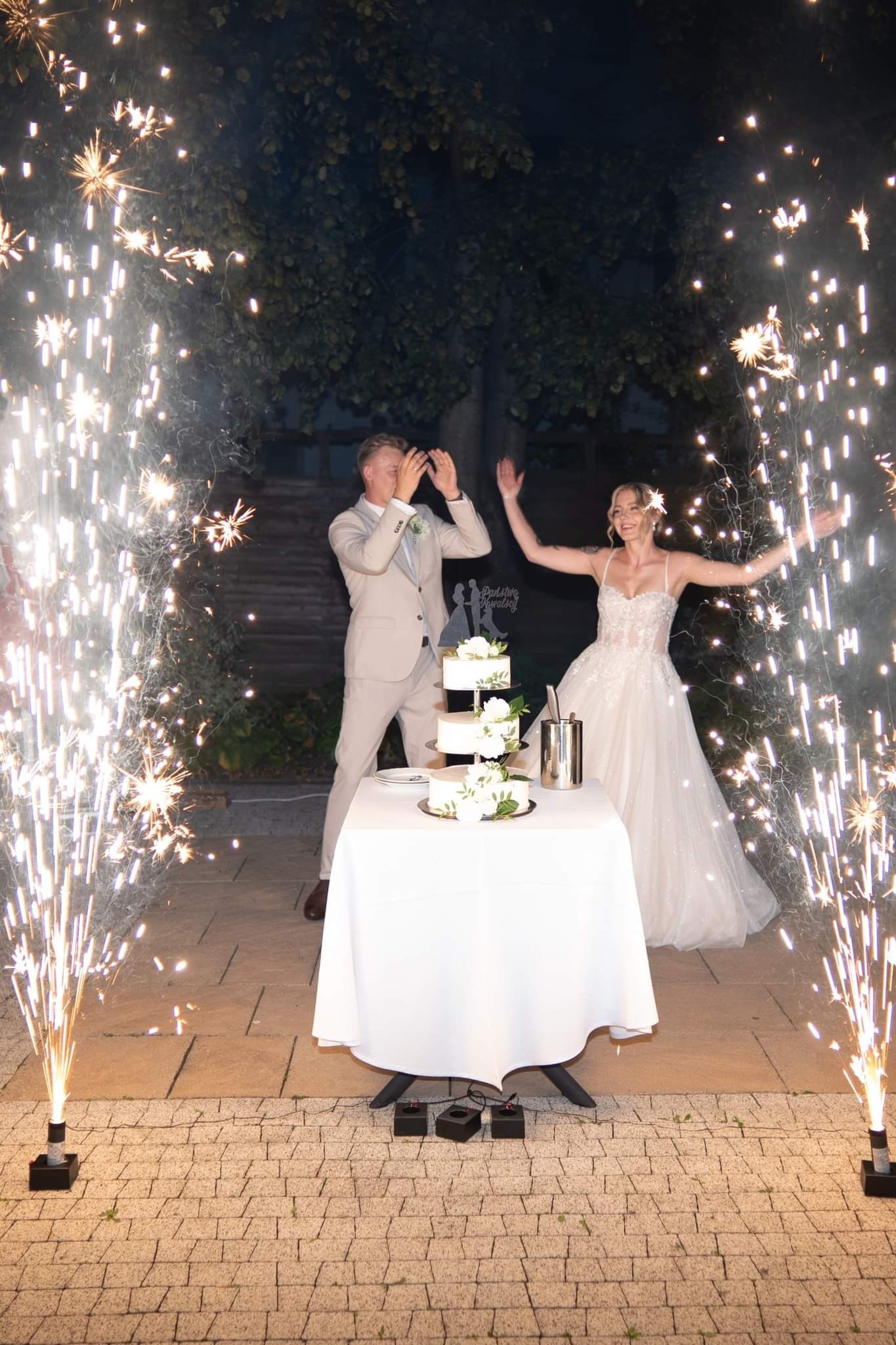 Nowożeńcy świętują tort weselny otoczeni fontannami iskier, na zewnątrz, nocą.