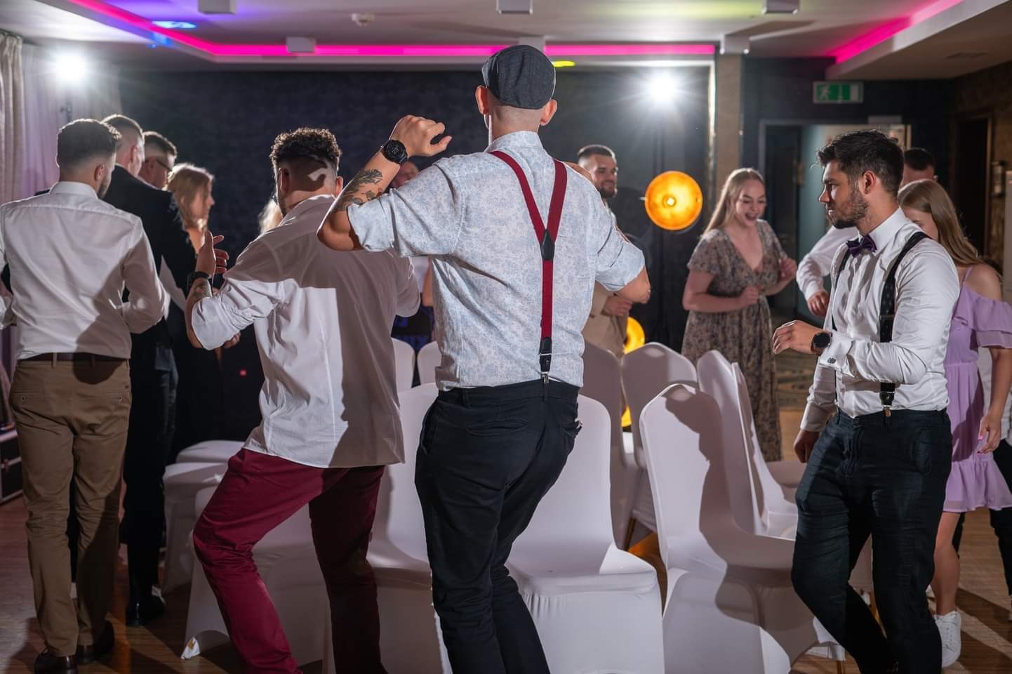 Grupa gości weselnych tańczy na parkiecie podczas przyjęcia, oświetlonym kolorowymi światłami, widoczne krzesła w tle.