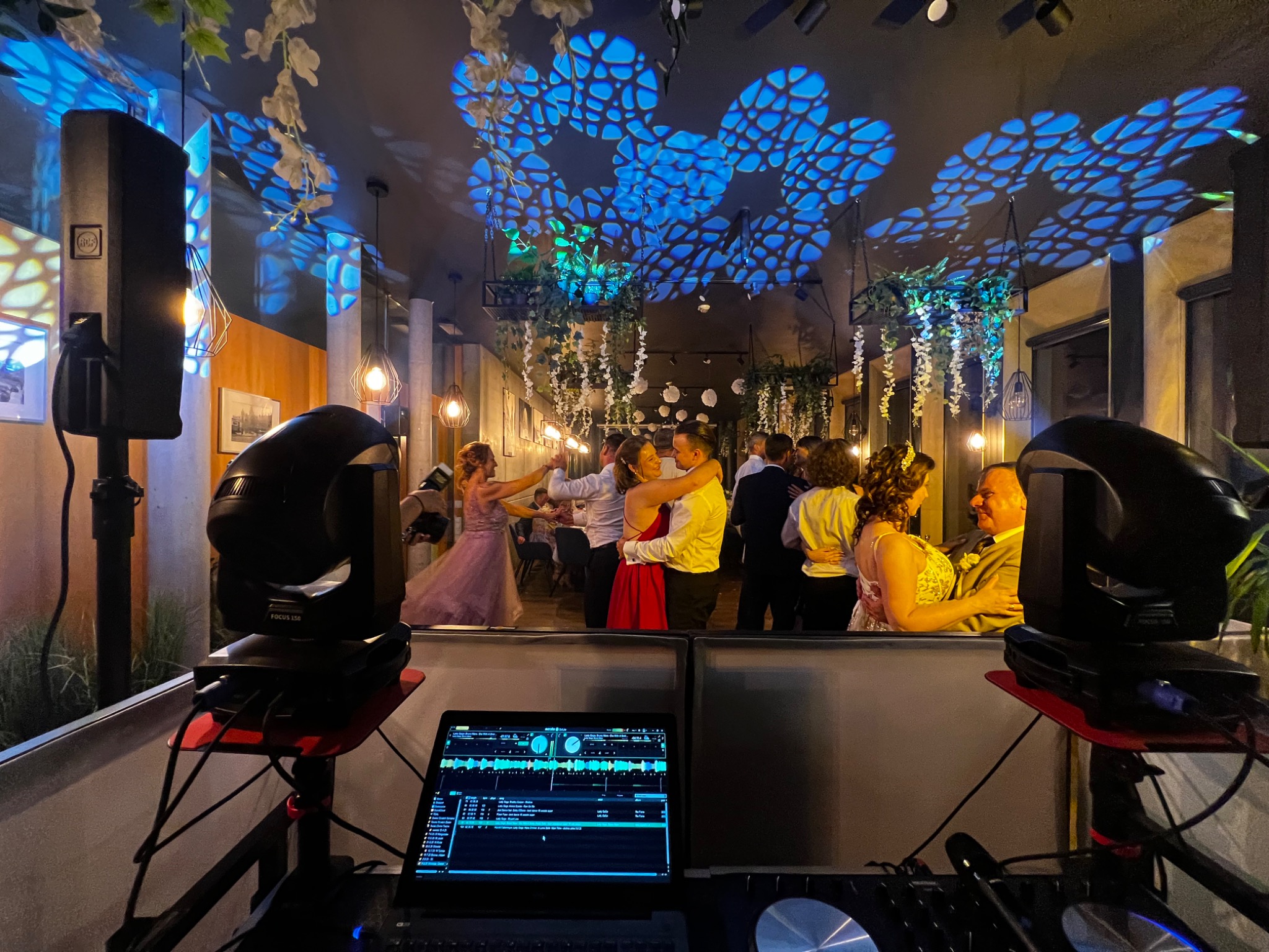 Parkiet taneczny z bawiącymi się ludźmi, oświetlony dekoracyjnymi światłami i nagłośniony sprzętem DJ-skim. Widoczne reflektory i laptop z programem muzycznym.