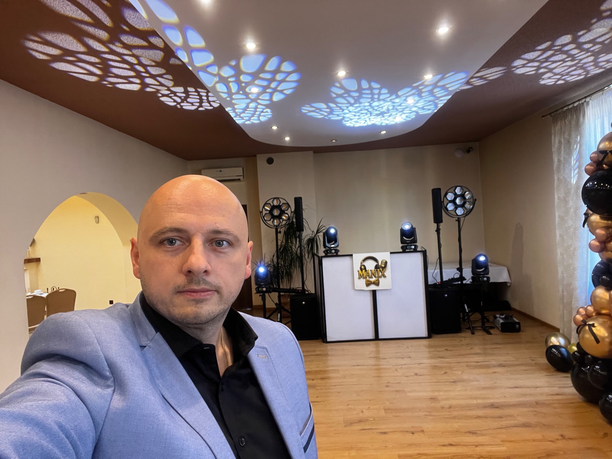 DJ w niebieskiej marynarce na tle sprzętu nagłośnieniowego i dekoracji balonowych, oświetlenie z efektem wzorów na suficie.