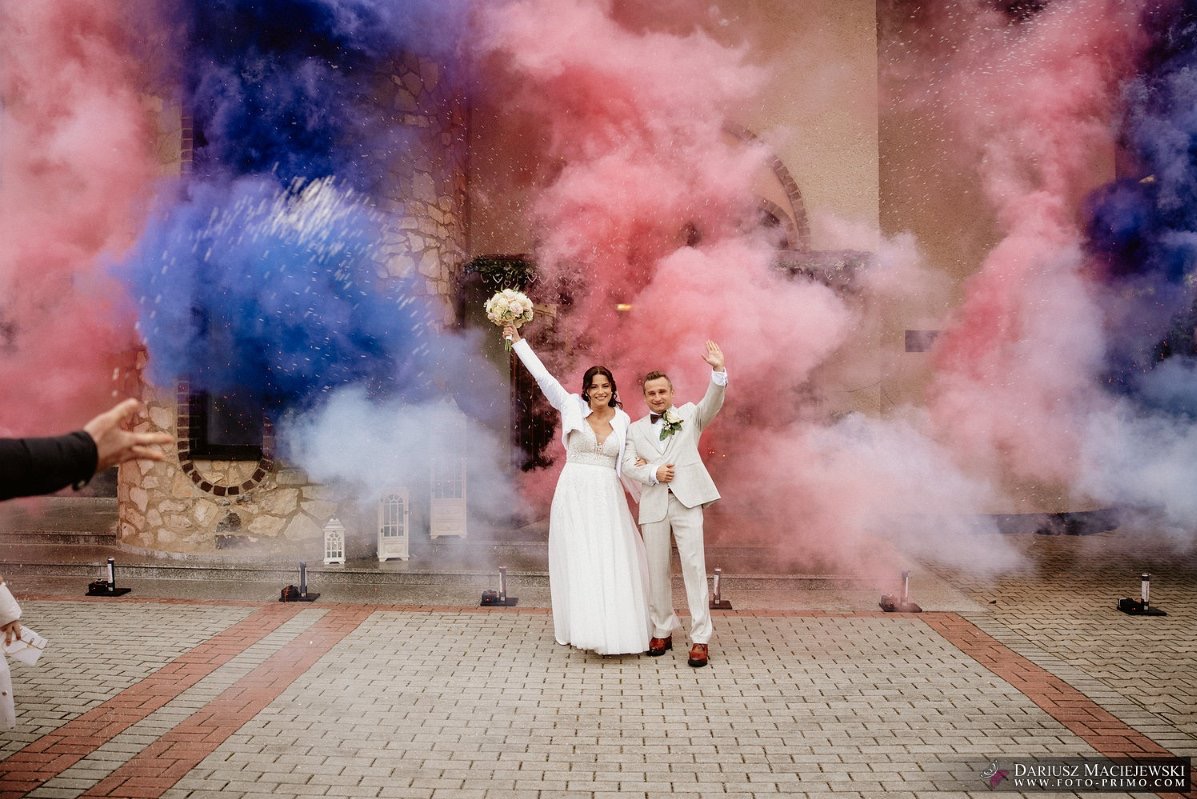 Uśmiechnięta para młoda w otoczeniu kolorowych dymów (różowy, niebieski) przed budynkiem. Panna młoda trzyma bukiet, oboje wznoszą ręce w geście radości.