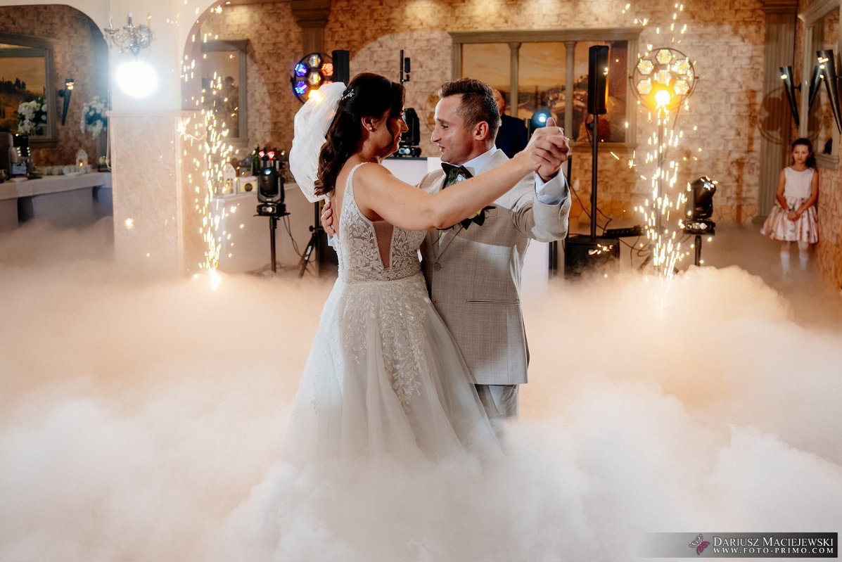 Para młoda tańczy w dymie na sali weselnej, oświetlenie sceniczne i fontanny iskier tworzą magiczną atmosferę. Dziewczynka w tle obserwuje.