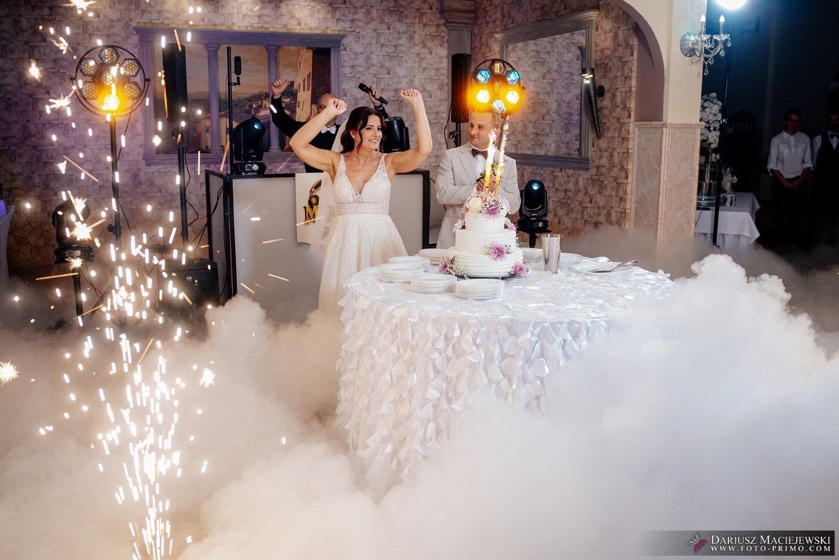 Uśmiechnięta para młoda przy torcie weselnym, w tle DJ i efekty specjalne: światła, dym i iskry. Dekoracja sali w jasnych barwach.
