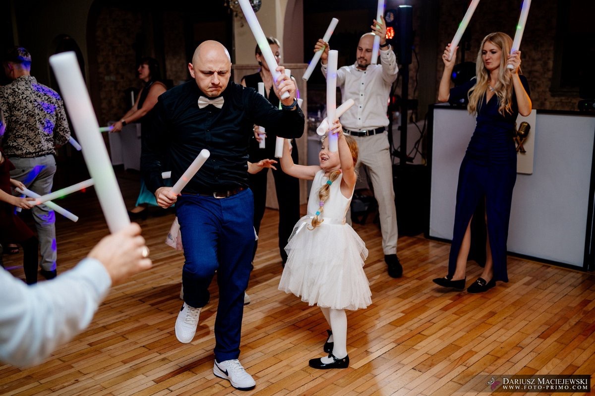 Uczestnicy imprezy, w tym dziecko w białej sukience, tańczą z podświetlanymi pałkami na parkiecie. W tle DJ-ka i inni goście.