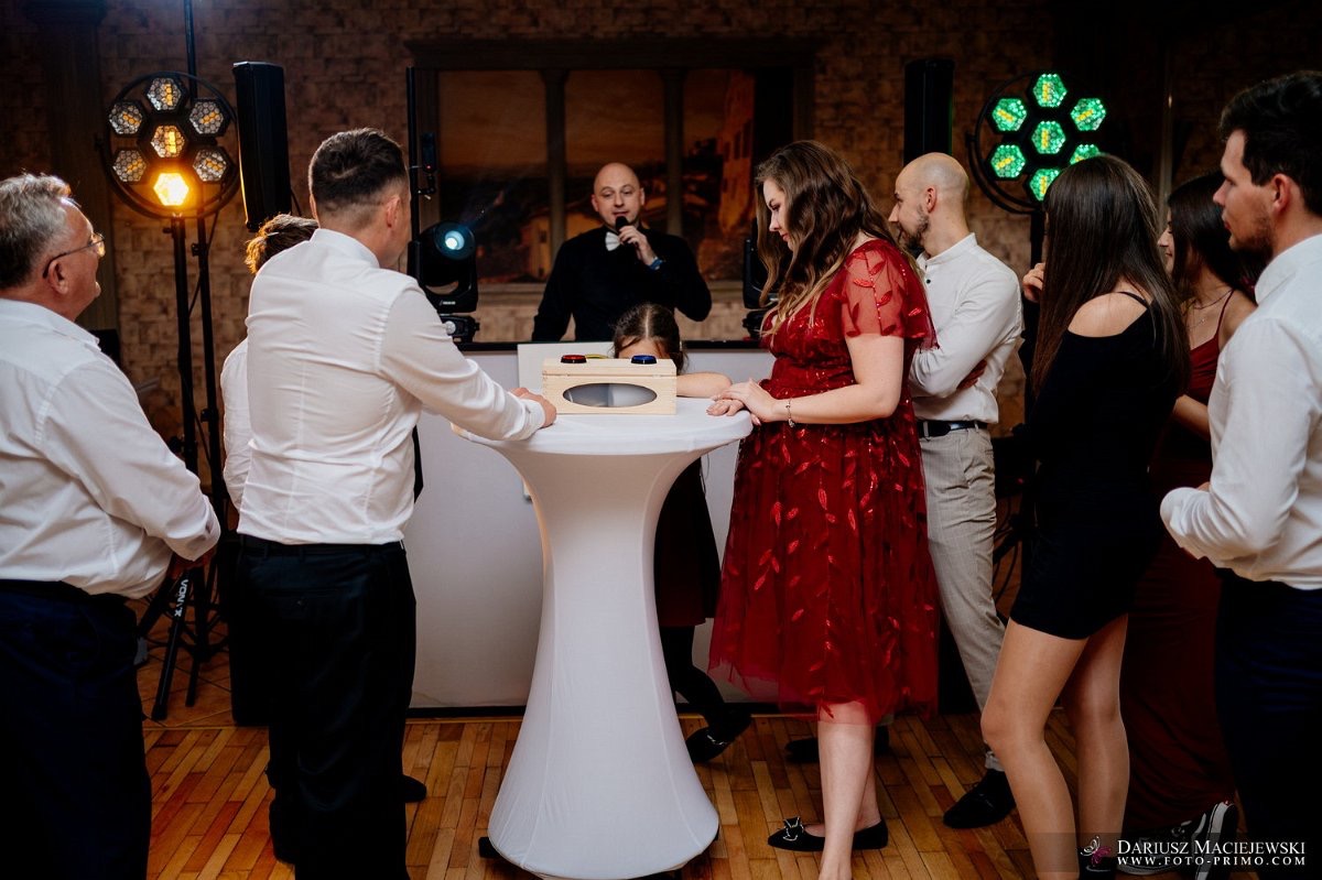 Event z wodzirejem: goście w eleganckich strojach obserwują interaktywną zabawę z drewnianą skrzynką na białym stoliku, oświetlenie sceniczne w tle.