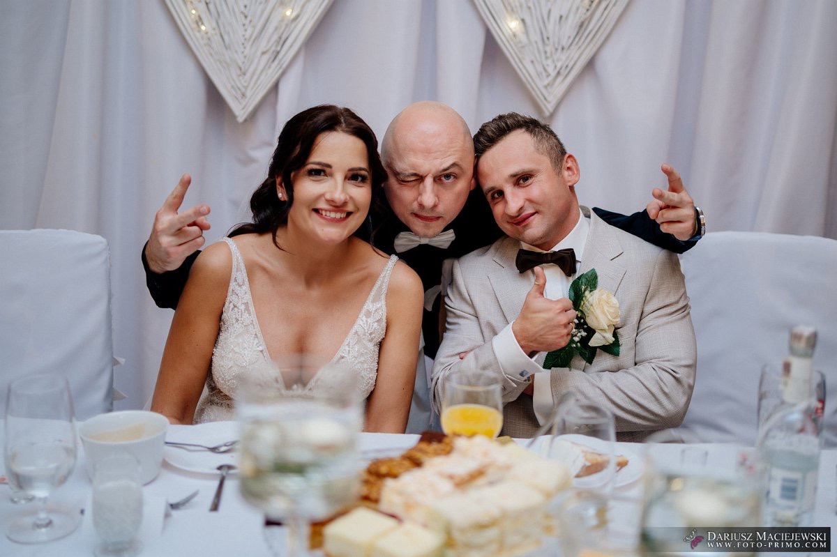 Uśmiechnięta panna młoda w koronkowej sukni, pan młody w beżowym garniturze z butonierką i mężczyzna w muszce pozują do zdjęcia przy stole weselnym.