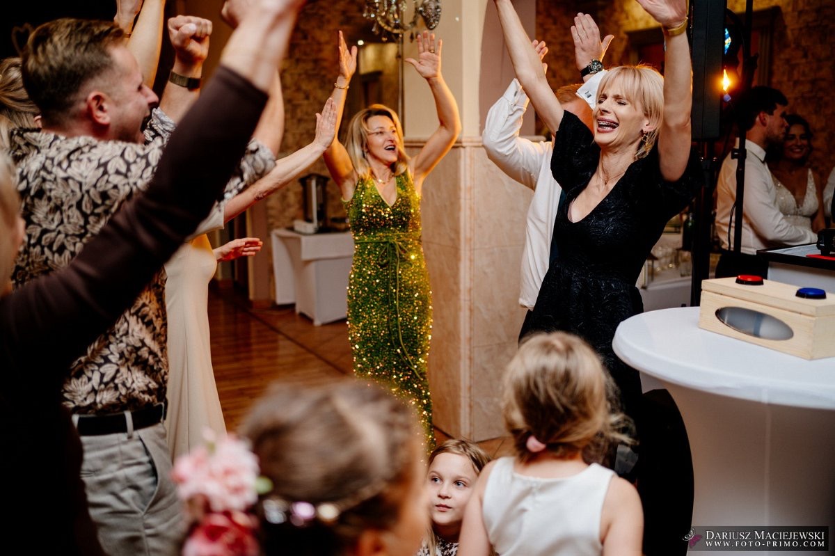 Rozbawieni goście weselni z podniesionymi rękami, w tle para młoda. Kobieta w zielonej sukni w cekiny i kobieta w czarnej sukience. Radosna atmosfera.