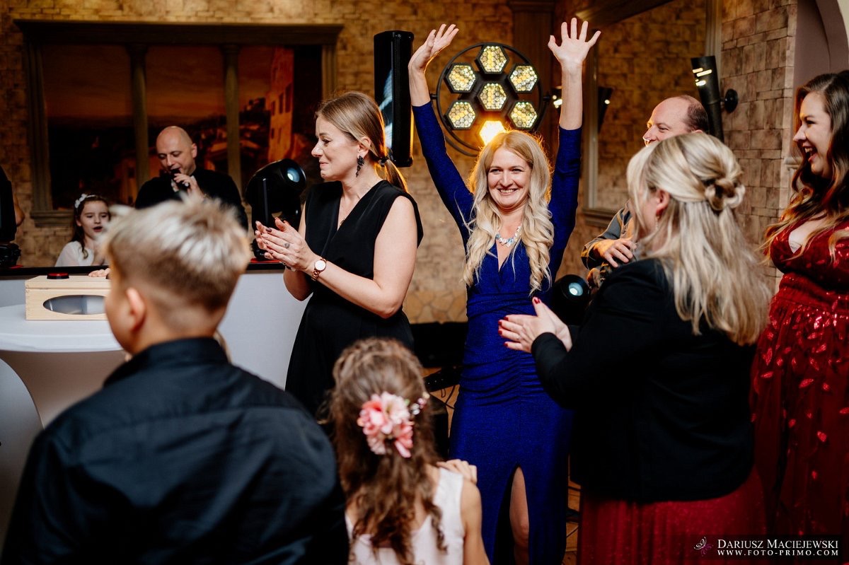 Uczestnicy imprezy w eleganckich strojach, w tle oświetlenie sceniczne i osoba z mikrofonem, tworzą radosną atmosferę podczas wydarzenia.