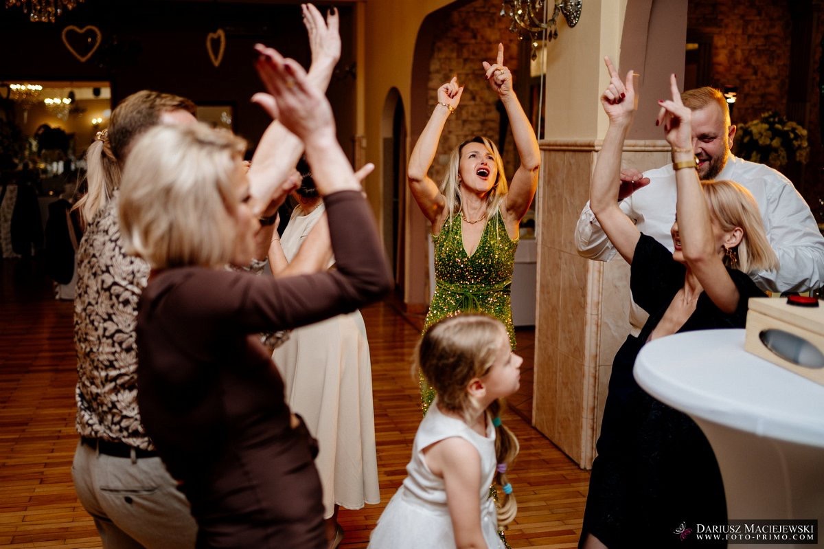 Rozbawieni goście weselni w tańcu, z podniesionymi rękami, w ciepłym oświetleniu sali balowej z parkietem i dekoracjami. Ujęcie z perspektywy parkietu.