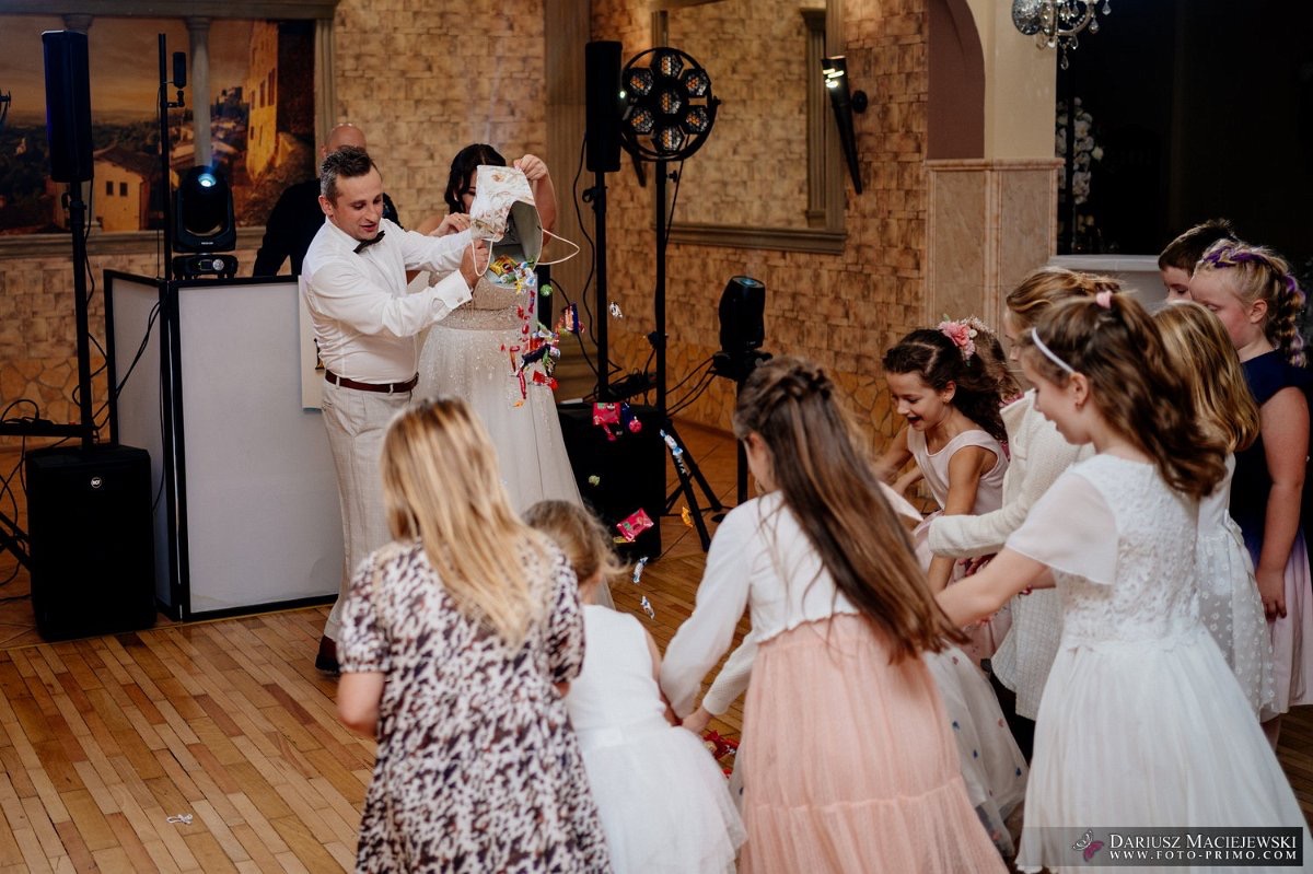 Para Młoda podczas oczepin rzuca słodycze do tańczących dzieci na sali weselnej. W tle sprzęt nagłośnieniowy i dekoracja ściany.