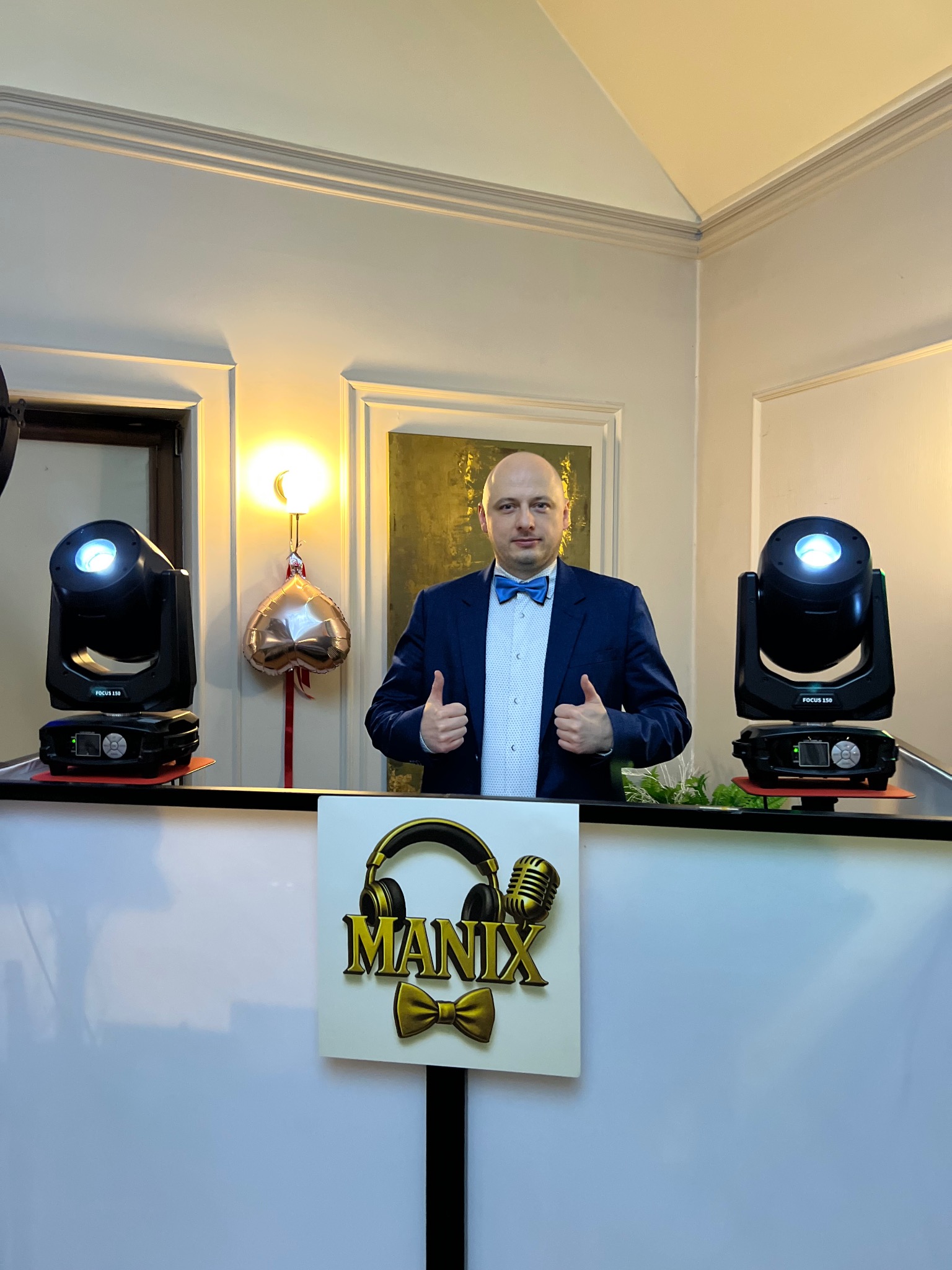 DJ w niebieskiej muszce za konsoletą z logo 'MANIX', oświetlony reflektorami. Uśmiechnięty mężczyzna z uniesionymi kciukami, gotowy do występu na imprezie.