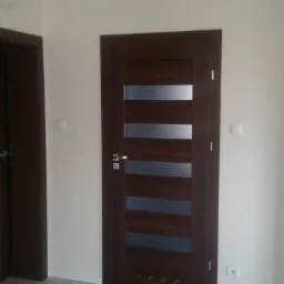 Dwie ciemnobrązowe, nowoczesne drzwi wewnętrzne z przeszkleniami i poziomymi panelami na tle świeżo pomalowanej, jasnej ściany.