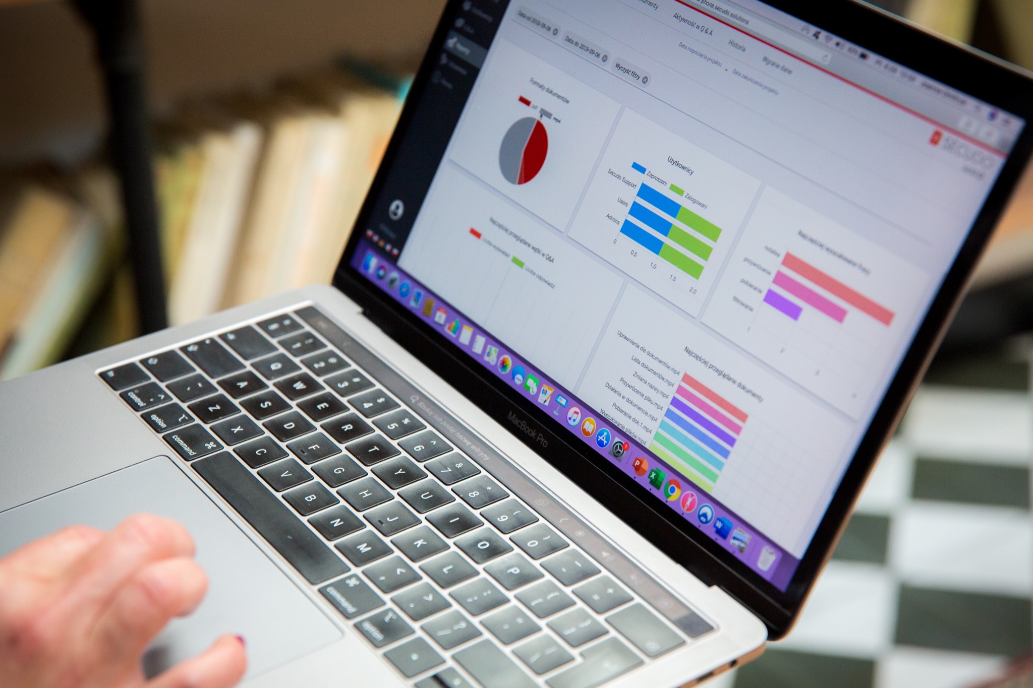 Laptop MacBook Pro wyświetlający wykresy i dane analityczne, prawdopodobnie związane z zarządzaniem dokumentami, na tle regału z książkami.