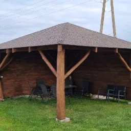 Drewniana altana z brązowym dachem krytym gontem bitumicznym, wyposażona w czarne krzesła i stolik, na trawniku, z grillem i huśtawką w tle.